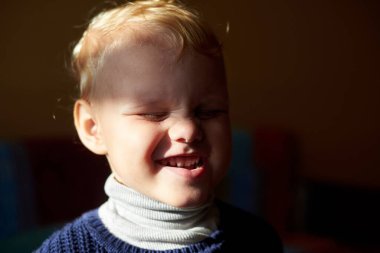A three-year-old blonde boy squints against the sun, grimaces and smiles at home. clipart