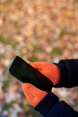 Turuncu eldivenli bir kadının elleri sonbahar arka planında bir akıllı telefon tutar.