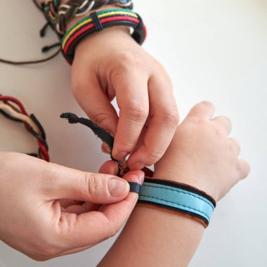 Kadın, beyaz arka planda bir çocuğun koluna deri bir bilezik bağlıyor..