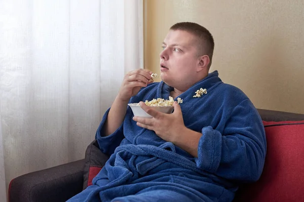 Fat man in a robe eating popcorn while sitting on the couch, obesity and improper lifestyle concept.