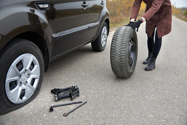 A young woman rolls spare tire near her car with a flat tire, trouble on the road.