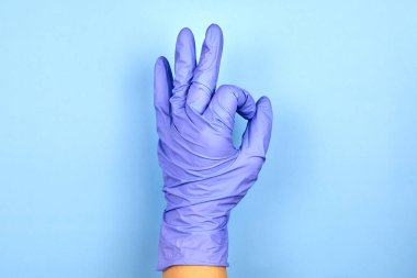 Tıbbi eldiven giymiş bir kadın mavi arka planda bir jest olduğunu gösteriyor..