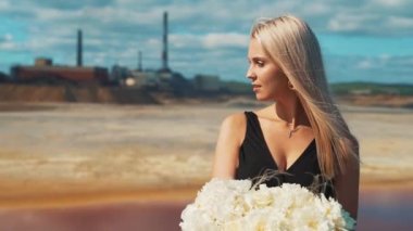 Siyah elbiseli genç bir sarışın kız ellerinde çiçekler ve arka planda sigara fabrikasının bacalarında bir tıbbi maske..