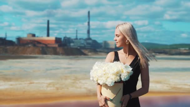 Una joven rubia con un vestido negro, flores en las manos y una máscara médica en el fondo de las chimeneas de una fábrica de ahumado de una planta metalúrgica. — Vídeos de Stock