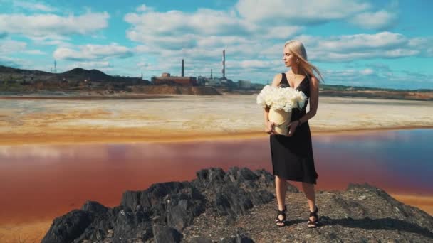 Een jong blond meisje in een zwarte jurk, bloemen in haar handen en een medisch masker op de achtergrond van de rookfabriek schoorstenen van een metallurgische plant. — Stockvideo
