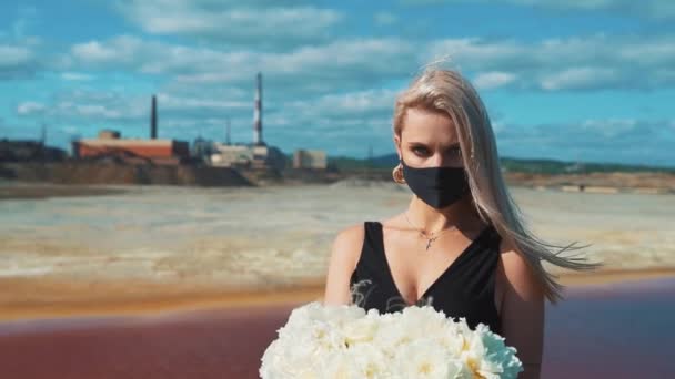 Seorang gadis muda berambut pirang dalam gaun hitam, bunga di tangannya dan topeng medis di latar belakang cerobong pabrik merokok dari tanaman metalurgi. — Stok Video
