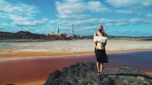 Una joven rubia con un vestido negro, flores en las manos y una máscara médica en el fondo de las chimeneas de una fábrica de ahumado de una planta metalúrgica. — Vídeos de Stock