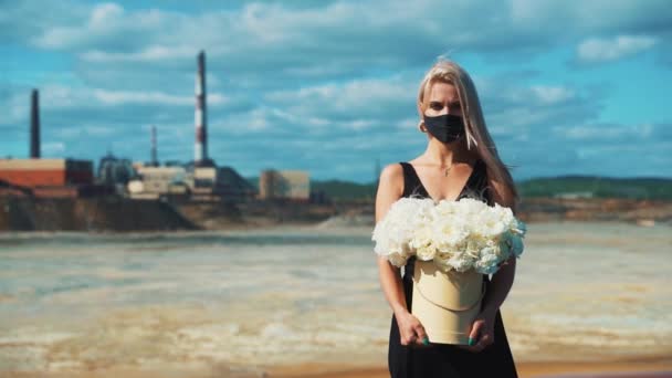 Młoda blondynka w czarnej sukience, kwiaty w dłoniach i maska medyczna na tle kominów fabryki dymu metalurgicznego. — Wideo stockowe