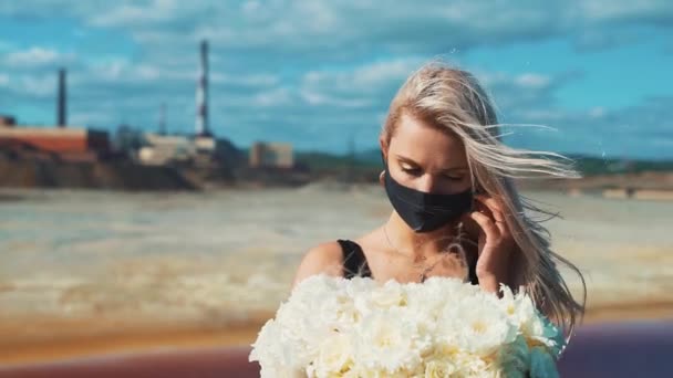 Una joven rubia con un vestido negro, flores en las manos y una máscara médica en el fondo de las chimeneas de una fábrica de ahumado de una planta metalúrgica. — Vídeos de Stock