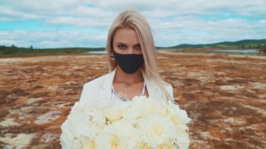 Elinde şakayık demeti olan beyaz takım elbiseli bir kadın. Yanmış bir tarlanın ortasında duruyor, siyah bir tıbbi maske takıyor. Hava kirliliği, enfeksiyon, salgın