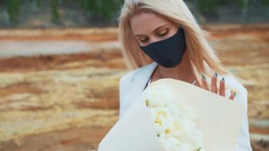 Elinde şakayık demeti olan beyaz takım elbiseli bir kadın. Yanmış bir tarlanın ortasında duruyor, siyah bir tıbbi maske takıyor. Hava kirliliği, enfeksiyon, salgın