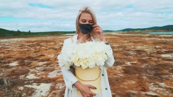 Μια γυναίκα με λευκό κοστούμι με ένα μπουκέτο παιώνιες στα χέρια της. Στέκεται στη μέση ενός καμένου αγρού, φορώντας μια μαύρη ιατρική μάσκα. Η έννοια της ατμοσφαιρικής ρύπανσης, μόλυνσης, πανδημίας — Αρχείο Βίντεο