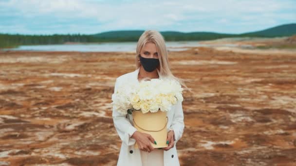 Una mujer con un traje blanco con un ramo de peonías en sus manos. Está en medio de un campo quemado, con una máscara médica negra. El concepto de contaminación atmosférica, infección, pandemia — Vídeos de Stock