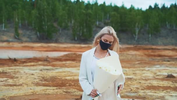 Een vrouw in een wit pak met een boeket pioenrozen in haar handen. Ze staat midden in een verschroeid veld, met een zwart medisch masker op. Het concept van luchtverontreiniging, infectie, pandemie — Stockvideo