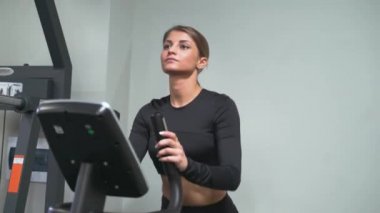 Spor salonundaki esmer bir kadın siyah eşofman ve spor ayakkabı giyen bir ayakçıyla antrenman yapıyor. Kilo vermek için aerobik kardiyo egzersizi yapıyor. Yağ yanığı