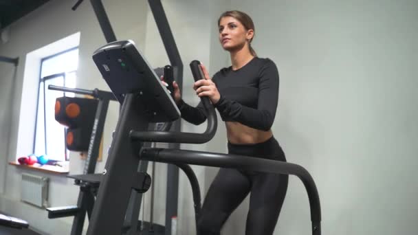 Een brunette in de sportschool traint op een stepper in een zwart trainingspak en sneakers. Voert aerobe cardiotraining uit voor gewichtsverlies. Vetverbranding — Stockvideo