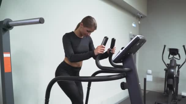 En brunett kvinna i gymmet tränar på en stepper i svart träningsoverall och sneakers. Utför aerob konditionsträning för viktminskning. Fettförbränning — Stockvideo