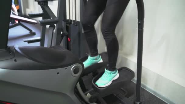 En brunett kvinna i gymmet tränar på en stepper i svart träningsoverall och sneakers. Utför aerob konditionsträning för viktminskning. Fettförbränning — Stockvideo