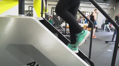 Jimnastik salonunda güzel bir esmer kız siyah spor giysisi ve spor ayakkabılarıyla stepper simülatörüyle antrenman yapıyor. Kilo vermek için aerobik kardiyo egzersizleri yapıyor. Yağ yanıyor. Merdivenlerden çık.