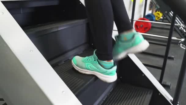 Une belle fille brune sportive dans la salle de gym s'entraîne sur un simulateur de stepper dans une tenue de sport noire et des baskets. Effectuer des exercices de cardio aérobie pour la perte de poids. Brûlure de graisse. Montez les escaliers. — Video