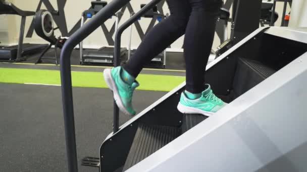Jimnastik salonunda güzel bir esmer kız siyah spor giysisi ve spor ayakkabılarıyla stepper simülatörüyle antrenman yapıyor. Kilo vermek için aerobik kardiyo egzersizleri yapıyor. Yağ yanıyor. Merdivenlerden çık. — Stok video