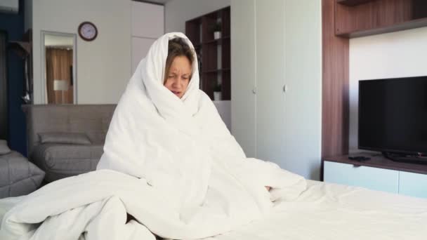 Síntomas de resfriado común. Una mujer joven enferma, sintiendo fiebre fría, se congela sin calefacción central, sintiendo frío en casa. Cubierto con una manta blanca cálida. Temblando, sentado en el sofá, estresado — Vídeo de stock