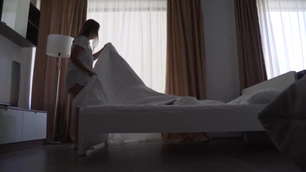 Mulher loira feliz faz a cama no quarto, espalha cobertor branco na cama. Levante e ventile o cobertor. Roupa de cama fresca, quarto limpo. Cama com travesseiro, edredão quente e lençóis de algodão branco — Vídeo de Stock