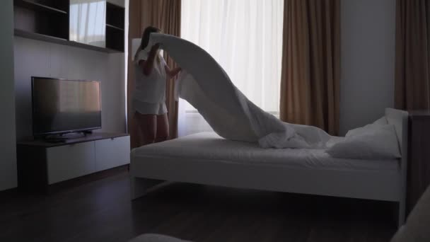Mujer rubia feliz hace la cama en el dormitorio, se extiende manta blanca en la cama. Levanta y ventila la manta. Ropa de cama fresca, dormitorio limpio. Cama con almohada, edredón caliente y sábanas de algodón blanco — Vídeo de stock