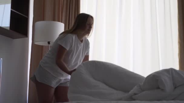 Mulher loira feliz faz a cama no quarto, espalha cobertor branco na cama. Levante e ventile o cobertor. Roupa de cama fresca, quarto limpo. Cama com travesseiro, edredão quente e lençóis de algodão branco — Vídeo de Stock