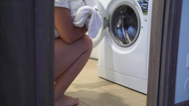Ev yapımı kıyafetleri olan genç bir kadın çamaşır makinesinin önünde oturur ve kirli beyaz şeyler yükler. Kadınların elleri çamaşır makinesine beyaz şeyler koyar. Çamaşır hazırlığı — Stok video