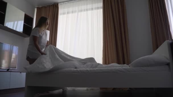 Lykkelig blondine kvinde gør sengen i soveværelset, spreder hvidt tæppe på sengen. Løft og ventiler tæppet. Frisk sengelinned, rent soveværelse. Seng med pude, varm dyne og hvidt bomuldssengetøj – Stock-video