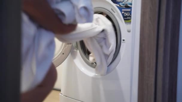 Młoda kobieta w domowych ciuchach siedzi przed pralką i ładuje brudne białe rzeczy. Kobiece ręce wkładają białe rzeczy do pralki. Przygotowanie prania — Wideo stockowe