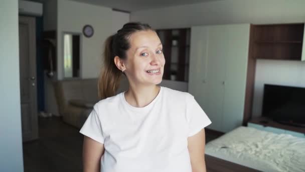 Glada kvinna med tandställning står ensam hemma i vit t-shirt och ler brett. Tandläkare, tandställning, behandling, leende koncept. Tandvård för friskt leende. Ortodontisk behandlingsplats — Stockvideo