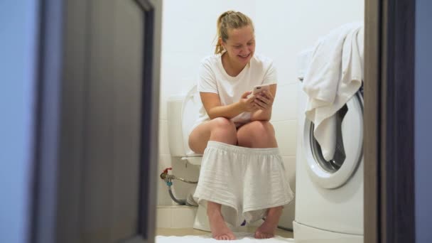 Mulher loira bonita feliz em pijama sorri e mantém o smartphone nas mãos, sentado no banheiro. Conversando e jogos no gadget. Estilo de vida com telefone. Usando smartphone na vida cotidiana — Vídeo de Stock