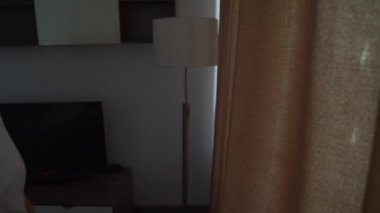 Mutlu, iyimser, beyaz tişörtlü genç bir kadın, rahat dairesini aydınlatmak için sabah güneşinin bej perdelerini açıyor. Pencereden tek başına bakıyor ve şehrin güzel manzarasından zevk alıyor.