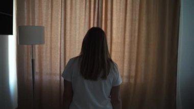 Mutlu, iyimser, beyaz tişörtlü genç bir kadın, rahat dairesini aydınlatmak için sabah güneşinin bej perdelerini açıyor. Pencereden tek başına bakıyor ve şehrin güzel manzarasından zevk alıyor.