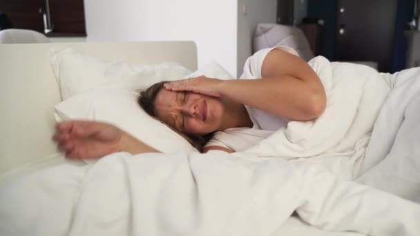 Jonge geïrriteerde vrouw kan niet slapen van hoofdpijn. Ligt alleen in bed in de slaapkamer onder een witte deken op een kussen in een witte pyjama en masseert haar slapen voor migraine. Slaapstoornis, slapeloosheid — Stockvideo