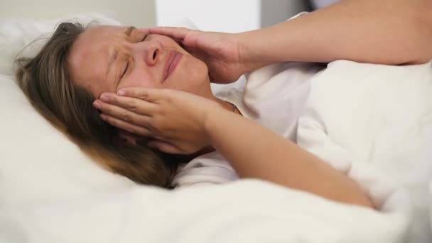 Jeune femme irritée ne peut pas dormir de maux de tête. Allongée seule au lit dans la chambre sous une couverture blanche sur un oreiller en pyjama blanc et massant ses tempes contre les migraines. Trouble du sommeil, insomnie — Video