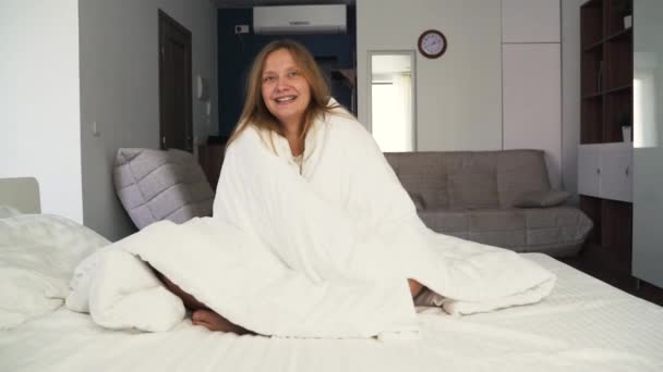 Vrolijke vrolijke jonge vrouw lacht en voelt zich zorgeloos verpakt in deken. Lachend en lachend om de camera die 's morgens in de slaapkamer op het bed zit. Ziet er gelukkig uit. Positieve emoties en gelach — Stockvideo