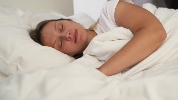 A jovem acorda de manhã cedo na cama. Mentiras, alongamentos e sorrisos. Goza de manhã cedo no quarto brilhante. Parece feliz deitado na almofada debaixo dos lençóis. covid-19, pandemia, auto-isolamento — Vídeo de Stock