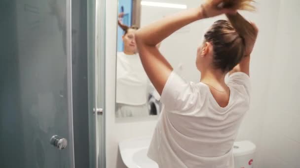 Happy beautiful blonde woman puts elastic band for long hair, makes ponytail and smiles, looking in the mirror in a bright bathroom. Pulls smooth hair into ponytail. Back view. Mirror reflection. — Stock Video