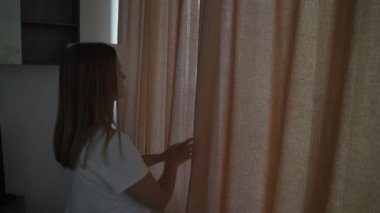 Mutlu, iyimser, beyaz tişörtlü genç bir kadın, rahat dairesini aydınlatmak için sabah güneşinin bej perdelerini açıyor. Pencereden tek başına bakıyor ve şehrin güzel manzarasından zevk alıyor.