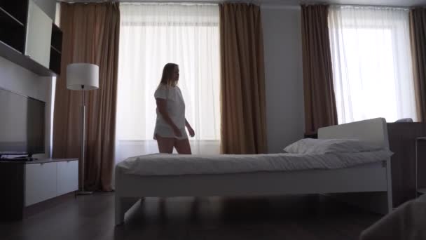 Een vermoeide, droevige vrouw in een wit T-shirt en korte broek valt met haar gezicht op het kussen op het bed bij het raam en valt in slaap. Ze voelt zich gestrest. Ontspan na een zware dag in een lichte slaapkamer. Alleen thuis. — Stockvideo