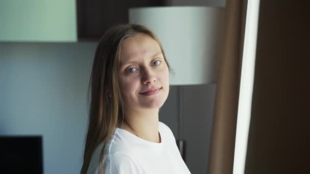 Une jeune femme en T-shirt blanc se tient près de la fenêtre seule à la maison et sourit. Pas de maquillage, cheveux lâches, boutons sur le visage. L'acné. Beauté naturelle — Video