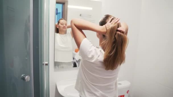 Mujer rubia hermosa feliz pone banda elástica para el pelo largo, hace cola de caballo y sonríe, mirando en el espejo en un baño luminoso. Tire del pelo liso en la cola de caballo. Vista trasera. Reflejo espejo. — Vídeo de stock