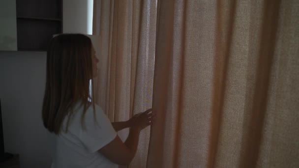 Glückliche optimistische junge Frau in weißem T-Shirt, die die beigefarbenen Vorhänge für die Morgensonne ablegt, um ihre gemütliche Wohnung zu erhellen. Sie blickt allein aus dem Fenster und genießt die schöne Aussicht auf das Stadtbild — Stockvideo