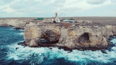 Aerial view of Cape Tarkhankut, Black Sea, Crimean peninsula. Blue stormy waves crash against rocky cliffs, water foams near the coast. Rocks with arches. Church on the cape. Overcast sky