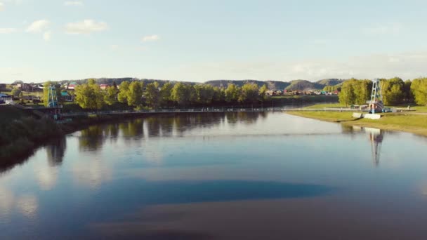 Vue aérienne de la passerelle au-dessus de la rivière, Oural Sud, Russie. Voler sur un drone au-dessus d'une large rivière sur les rives de laquelle une végétation dense et verte. Le soleil brille dans le ciel bleu et les nuages bougent — Video