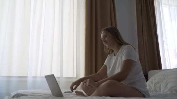 La mujer de camiseta blanca se sienta en la cama con auriculares y trabaja en un portátil. Comunicarse en videoconferencia en el ordenador, sonriendo y saludando con la mano. El concepto de trabajo desde casa, cuarentena — Vídeos de Stock