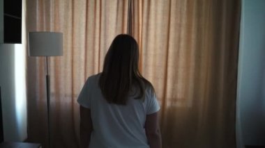 Mutlu, iyimser, beyaz tişörtlü genç bir kadın, rahat dairesini aydınlatmak için sabah güneşinin bej perdelerini açıyor. Pencereden tek başına bakıyor ve şehrin güzel manzarasından zevk alıyor.